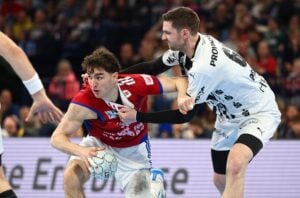 Zwei männliche Handballspieler liefern sich einen intensiven Wettkampf; der eine in einem roten Trikot hält den Ball fest und versucht, nach vorne zu kommen, während ein Spieler in einem weißen Trikot ihn verteidigt, indem er zum Block greift. Im Hintergrund ist ein unscharfes Publikum zu sehen.
