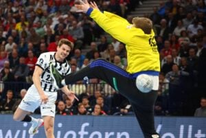 Ein Handballspieler in einem weißen Trikot springt, um den Ball zu schießen, während ein Torwart in Gelb sich ausstreckt, um das Tor zu verhindern. Im Hintergrund sieht eine Gruppe von Zuschauern zu.