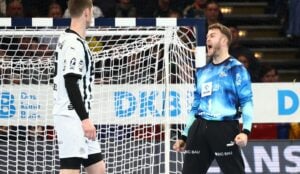 Ein Handballtorwart in einem blauen Trikot ballt die Fäuste und jubelt, während ein Spieler in einem schwarz-weißen Trikot in der Nähe vor dem Tornetz steht. Im Hintergrund sind Zuschauer zu sehen.