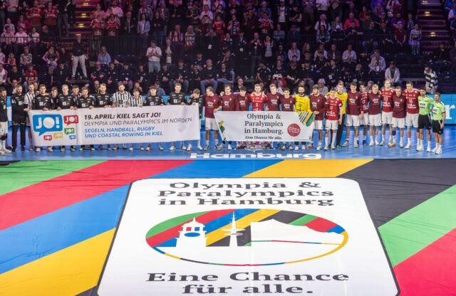 Zwei Handballmannschaften und Schiedsrichter stehen zusammen in einer Arena und halten Transparente, die für die Olympischen Spiele und Paralympics in Hamburg werben. Auf einem großen Bodenbanner steht: Olympia & Paralympics in Hamburg. Eine Chance für alle.