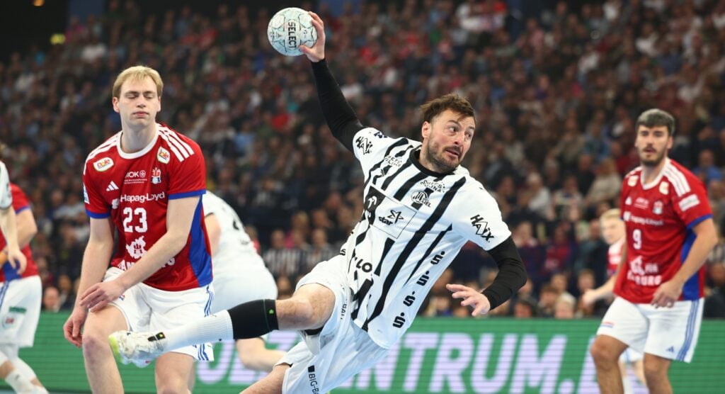 Handball 25/26 - HSV Hamburg Ein Handballspieler in einem schwarz-weißen Trikot springt, um den Ball zu werfen, während ein Verteidiger in einem rot-blauen Trikot in der Nähe steht; im Hintergrund ist eine überfüllte Arena zu sehen.
