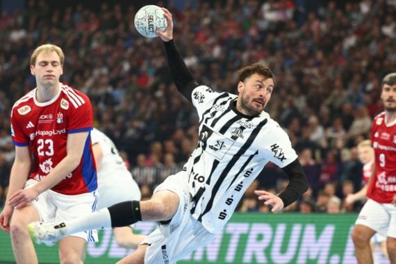 Ein Handballspieler in einem schwarz-weißen Trikot springt, um den Ball zu werfen, während ein Verteidiger in einem rot-blauen Trikot in der Nähe steht; im Hintergrund ist eine überfüllte Arena zu sehen.