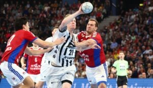Zwei Handballspieler, einer in Weiß und einer in Rot, kämpfen während eines intensiven Spiels um die Kontrolle über den Ball, während Mannschaftskameraden und ein Schiedsrichter im Hintergrund inmitten einer großen, sitzenden Menschenmenge zusehen.