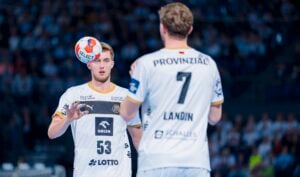 Zwei männliche Handballspieler in weißen Trikots, einer mit der Nummer 53 zur Kamera gewandt, der andere mit der Nummer 7 abgewandt. Der Spieler mit der Nummer 53 balanciert einen Handball in der Nähe seines Gesichts. Die Zuschauer sind im Hintergrund unscharf.