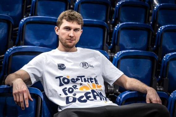 Ein Mann mit einem weißen "Together for Our Oceans"-T-Shirt sitzt entspannt in einer Reihe leerer blauer Stadionsitze und schaut in die Kamera.