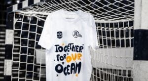 Ein weißes T-Shirt mit dem Aufdruck TOGETHER FOR OUR OCEAN in marineblauer und orangefarbener Schrift hängt an einem Fußballtornetz. Auf dem Shirt sind außerdem Logos des THW Kiel und einer anderen Organisation abgebildet.