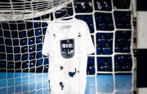 Ein weißes T-Shirt mit maritimen Motiven und einem großen Aufkleber mit der Aufschrift "Für die Küste" hängt an einem Fußballtornetz in einer leeren Sportarena.