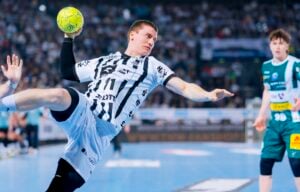 Ein männlicher Handballspieler in einem schwarz-weißen Trikot springt in der Luft und bereitet sich auf den Wurf eines gelben Balls vor. Ein anderer Spieler in Grün schaut zu, während eines Hallenspiels mit einer jubelnden Menge im Hintergrund.