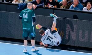 Ein Handballspieler in einem weißen Trikot fällt mit dem Ball in der Hand gegen eine Werbetafel, während ein Spieler in einem grünen Trikot in der Nähe steht. Die Zuschauer verfolgen das Geschehen von der Tribüne aus.