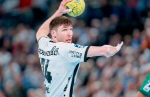 Ein männlicher Handballspieler in einem weißen Trikot bereitet sich darauf vor, einen gelben Ball während eines Spiels zu werfen. Der Hintergrund ist unscharf und deutet auf eine Halle mit Zuschauern hin.