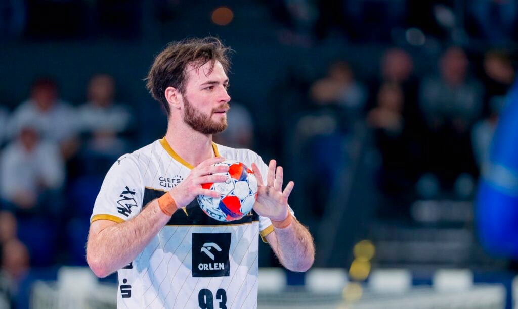 Ein männlicher Handballspieler in einem weiß-goldenen Trikot hält einen Ball mit beiden Händen und bereitet sich auf den Wurf vor, während im Hintergrund eine unscharfe Menschenmenge zu sehen ist.