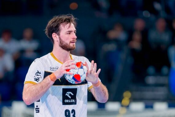 Ein männlicher Handballspieler in einem weiß-goldenen Trikot hält einen Ball mit beiden Händen und bereitet sich auf den Wurf vor, während im Hintergrund eine unscharfe Menschenmenge zu sehen ist.