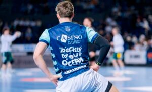 Ein Handballspieler steht mit den Händen in den Hüften auf einem Hallenplatz und trägt ein blaues Trikot mit der Aufschrift Together for our Ocean auf dem Rücken. Der Hintergrund ist unscharf und zeigt andere Spieler und Zuschauer.