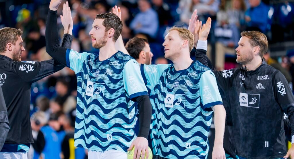 Drei männliche Athleten in blau gemusterten Trikots klatschen während eines Sportereignisses mit ihren Mannschaftskameraden ab, wobei die Zuschauer im unscharfen Hintergrund zu sehen sind. Alle wirken konzentriert und im Teamgeist engagiert.