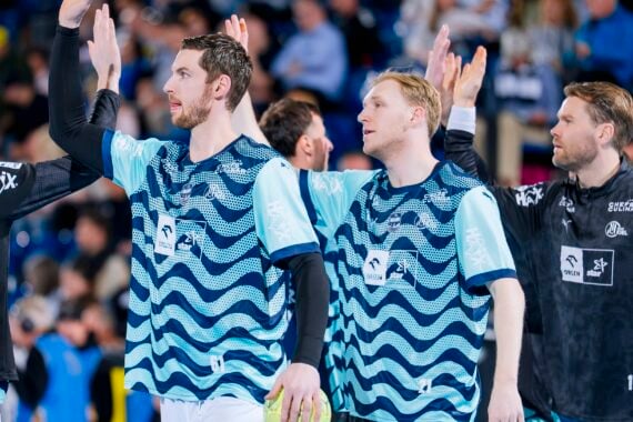 Drei männliche Athleten in blau gemusterten Trikots klatschen während eines Sportereignisses mit ihren Mannschaftskameraden ab, wobei die Zuschauer im unscharfen Hintergrund zu sehen sind. Alle wirken konzentriert und im Teamgeist engagiert.