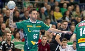 Ein Handballspieler im grünen Trikot mit der Nummer 67 bereitet sich darauf vor, den Ball während eines Spiels zu werfen. Im Hintergrund sind andere Spieler und Zuschauer zu sehen, die eine intensive Spielatmosphäre schaffen.
