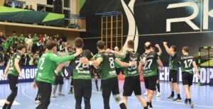 Eine Gruppe männlicher Handballspieler in grün-schwarzen Trikots feiert auf einem Hallenplatz vor jubelnden Fans auf der Tribüne. Die Stimmung ist ausgelassen, und die Mannschaft scheint ein Spiel gewonnen zu haben.
