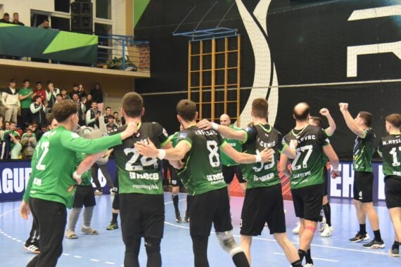 Eine Gruppe männlicher Handballspieler in grün-schwarzen Trikots feiert auf einem Hallenplatz vor jubelnden Fans auf der Tribüne. Die Stimmung ist ausgelassen, und die Mannschaft scheint ein Spiel gewonnen zu haben.