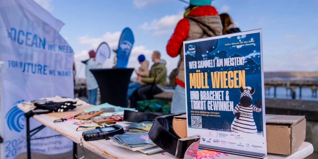 Nahaufnahme eines Tisches mit Müllsammelgeräten und einem Schild in deutscher Sprache zum Thema Müllsammeln, mit Menschen und Bannern zum Thema Meer im Hintergrund auf einem Pier in der Nähe des Wassers.