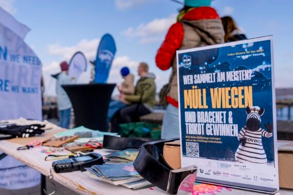 Nahaufnahme eines Tisches mit Müllsammelgeräten und einem Schild in deutscher Sprache zum Thema Müllsammeln, mit Menschen und Bannern zum Thema Meer im Hintergrund auf einem Pier in der Nähe des Wassers.