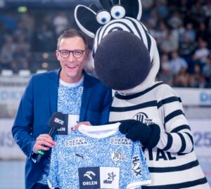 Ein lächelnder Mann mit Brille und blauem Blazer steht neben einer Person in einem Zebra-Maskottchen-Kostüm, beide halten ein THW Kiel-Trikot auf dem Boden einer Sporthalle. Der Mann hält ein Mikrofon in der Hand.