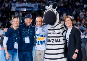 Vier Personen stehen lächelnd neben einem Zebra-Maskottchen bei einer Hallenveranstaltung der Handball-Bundesliga des THW Kiel. Lässig gekleidet mit Veranstaltungsplaketten zelebrieren sie den Teamgeist, während im Hintergrund die Zuschauer in der Halle jubeln.