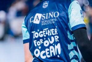 Eine Person trägt ein blaues Sporttrikot mit der Aufschrift CASINO SCHLESWIG HOLSTEIN und TOGETHER FOR OUR OCEAN! auf dem Rücken. Der Fokus liegt auf dem Rücken des Trikots, der Hintergrund ist unscharf.