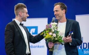 Zwei Männer stehen auf der Bühne; der eine im Smoking hält ein Mikrofon und berührt die Schulter des anderen, der lächelnd einen Blumenstrauß und eine Auszeichnung in der Hand hält. Im Hintergrund sind unscharfe Transparente zu sehen.