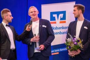 Drei Männer in förmlicher Kleidung stehen auf der Bühne; einer wird mit einem Mikrofon interviewt, während er eine Trophäe hält, ein anderer hält einen Blumenstrauß. Im Hintergrund ist ein großes Banner der Volksbanken Raiffeisenbanken zu sehen.