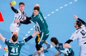 Ein Handballspiel in Aktion: Ein Spieler in einem weiß-schwarzen Trikot versucht einen Wurf, während er von Spielern in grünen Trikots verteidigt wird. Andere Spieler strecken die Hand aus oder bereiten sich darauf vor, ihn auf dem blauen Spielfeld abzufangen.