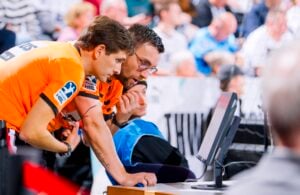 Zwei Schiedsrichter in orangefarbenen Hemden prüfen während eines Sportereignisses einen Monitor, wobei sich einer nach vorne lehnt und der andere auf den Bildschirm zeigt; im Hintergrund ist eine verschwommene Menschenmenge zu sehen.