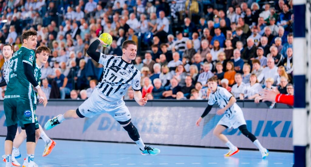 HANDBALL 2025/26: THW Kiel - ThSV Eisenach Ein Handballspieler in einem weiß-schwarz gestreiften Trikot springt in der Luft und versucht, den Ball in Richtung Tor zu werfen, während die Verteidiger in Grün und die Mitspieler zusehen. Eine große Menschenmenge füllt die Arena im Hintergrund.