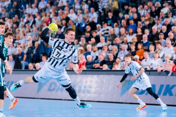 Ein Handballspieler in einem weiß-schwarz gestreiften Trikot springt in der Luft und versucht, den Ball in Richtung Tor zu werfen, während die Verteidiger in Grün und die Mitspieler zusehen. Eine große Menschenmenge füllt die Arena im Hintergrund.