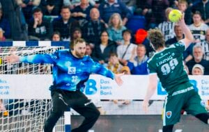 Ein Handballspieler im grünen Trikot bereitet sich im Sprung auf einen Schuss vor. Er steht einem Torwart im blauen Trikot gegenüber, der seine Arme weit ausstreckt, während im Hintergrund Zuschauer von der Tribüne aus zusehen.