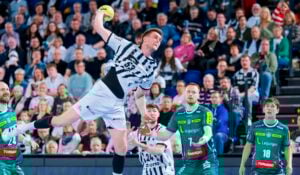 Ein Handballspieler in einem schwarz-weißen Trikot springt in der Luft, um den Ball zu werfen, während die gegnerischen Spieler in grünen Trikots abwehrend zusehen. Das Geschehen spielt sich in einer überfüllten Sporthalle ab.