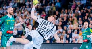 Ein männlicher Handballspieler in einem weiß-schwarzen Trikot springt, um den Ball zu werfen, während Spieler in grünen Trikots den Ball verteidigen. Die Szene spielt sich in einer vollbesetzten Halle ab, in der Zuschauer zuschauen.