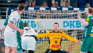 Ein Handballtorwart in einem gelben Trikot streckt seine Arme aus, um einen Schuss abzufangen, während Spieler in grün-weißen Trikots in der Nähe des Tores um die Wette spielen. Die Zuschauer verfolgen das Geschehen von der Tribüne im Hintergrund.