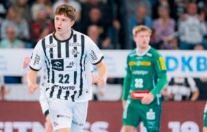 Ein männlicher Handballspieler in einem schwarz-weiß gestreiften Trikot mit der Nummer 22 jubelt auf dem Spielfeld, während ein anderer Spieler in einem grünen Trikot unscharf im Hintergrund steht. Auf der Tribüne sind Zuschauer zu sehen.