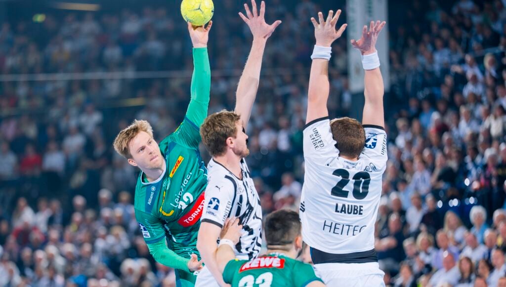 Ein männlicher Handballspieler in einem grünen Trikot springt, um den Ball zu werfen, während zwei gegnerische Spieler in weißen Trikots mit erhobenen Armen versuchen, ihn während eines Spiels in einer überfüllten Halle zu blockieren.