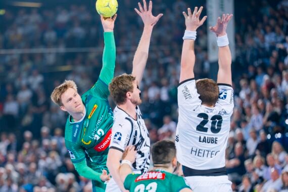 Ein männlicher Handballspieler in einem grünen Trikot springt, um den Ball zu werfen, während zwei gegnerische Spieler in weißen Trikots mit erhobenen Armen versuchen, ihn während eines Spiels in einer überfüllten Halle zu blockieren.