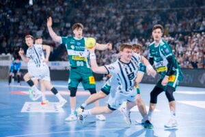 Ein Handballspieler in einem weiß-schwarzen Trikot wirft den Ball nach vorne, während die gegnerischen Spieler in Grün verteidigen. Andere Spieler und eine jubelnde Menge füllen die hell erleuchtete Halle.