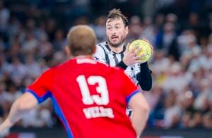 Ein Handballspieler in einem weiß-schwarzen Trikot hält den Ball und blickt nach vorne, während ein Spieler in einem roten Trikot mit der Nummer 13 und dem Namen Villar ihm auf einem überfüllten Hallenplatz gegenübersteht.