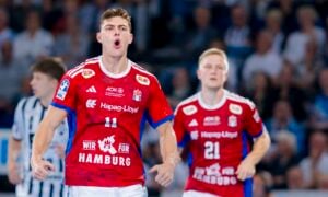 Ein männlicher Handballspieler in einem roten Trikot mit der Aufschrift Hamburg schreit während eines Spiels lautstark. Hinter ihm ist ein anderer Mannschaftskamerad in einem ähnlichen Trikot zu sehen, und der Hintergrund zeigt eine unscharfe Menge und Gegner.