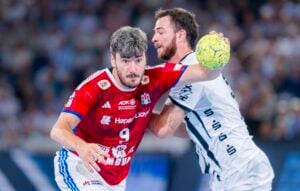 Zwei männliche Handballspieler kämpfen während eines Spiels um den Besitz eines gelben Balls. Der eine trägt ein rotes Trikot, der andere ein weißes Trikot. Im Hintergrund ist ein Publikum zu sehen.