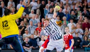 Ein Handballspieler in einem weiß-schwarzen Trikot springt, um den Ball auf das Tor zu schießen, während der Torwart in einem gelben Trikot sich darauf vorbereitet, den Ball zu blocken. Eine große Zuschauermenge verfolgt das intensive Spiel.