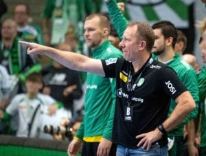 Ein Trainer in einem schwarzen Hemd zeigt und gibt Anweisungen während eines Handballspiels, während im Hintergrund Mannschaftsmitglieder in grünen Trikots und Fans zu sehen sind.