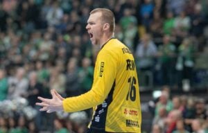 Ein Handballspieler in einem gelben Trikot mit der Nummer 16 schreit während eines Spiels leidenschaftlich und gestikuliert mit beiden Händen. Im Hintergrund ist die verschwommene Menge in Grün und Grau zu sehen.