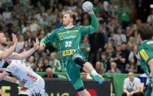 Ein männlicher Handballspieler in einem grünen Trikot springt in der Luft, um den Ball zu werfen. Er ist umgeben von weiß gekleideten Gegnern, während eines Spiels mit einer jubelnden Menge im Hintergrund.