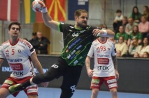 Ein männlicher Handballspieler in einem schwarz-grünen Trikot springt in die Luft, um den Ball zu werfen, während zwei gegnerische Spieler in weiß-roten Trikots den Ball verteidigen. Die Zuschauer schauen von den Tribünen im Hintergrund zu.