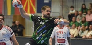 Ein männlicher Handballspieler in einem schwarz-grünen Trikot mit der Nummer 44 bereitet sich darauf vor, den Ball während eines Spiels zu werfen, während im Hintergrund Spieler in weißen Trikots und eine Zuschauermenge zusehen.